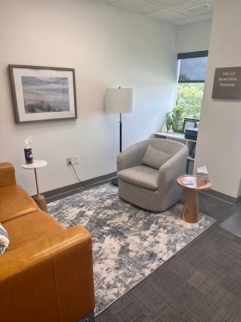 A cozy office space with a light brown leather couch, a gray armchair, and a small round wooden side table. A standing lamp, framed beach photo, and a sign saying "Hello Beautiful Person" decorate the room. There is a window revealing greenery outside.